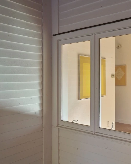 Interior view featuring a white-framed window, shuttered wall, and framed art, capturing a blend of architectural elements and artistic display.