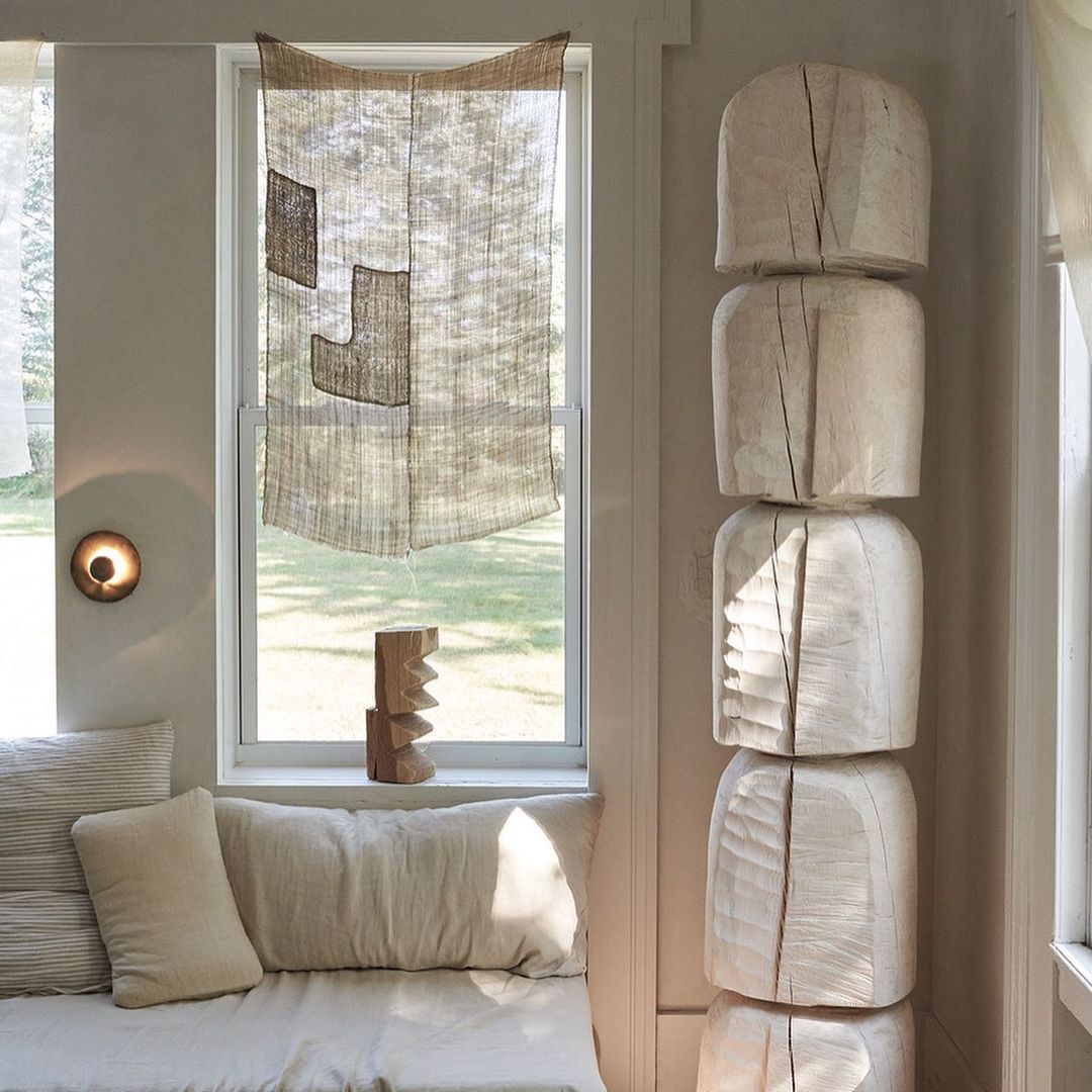 Living room interior with light beige textiles, pillows, and a geometric wood sculpture near a window with neutral, woven window covering.