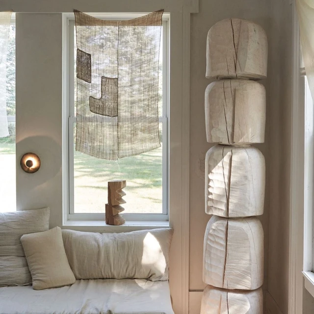 Living room interior with light beige textiles, pillows, and a geometric wood sculpture near a window with neutral, woven window covering.