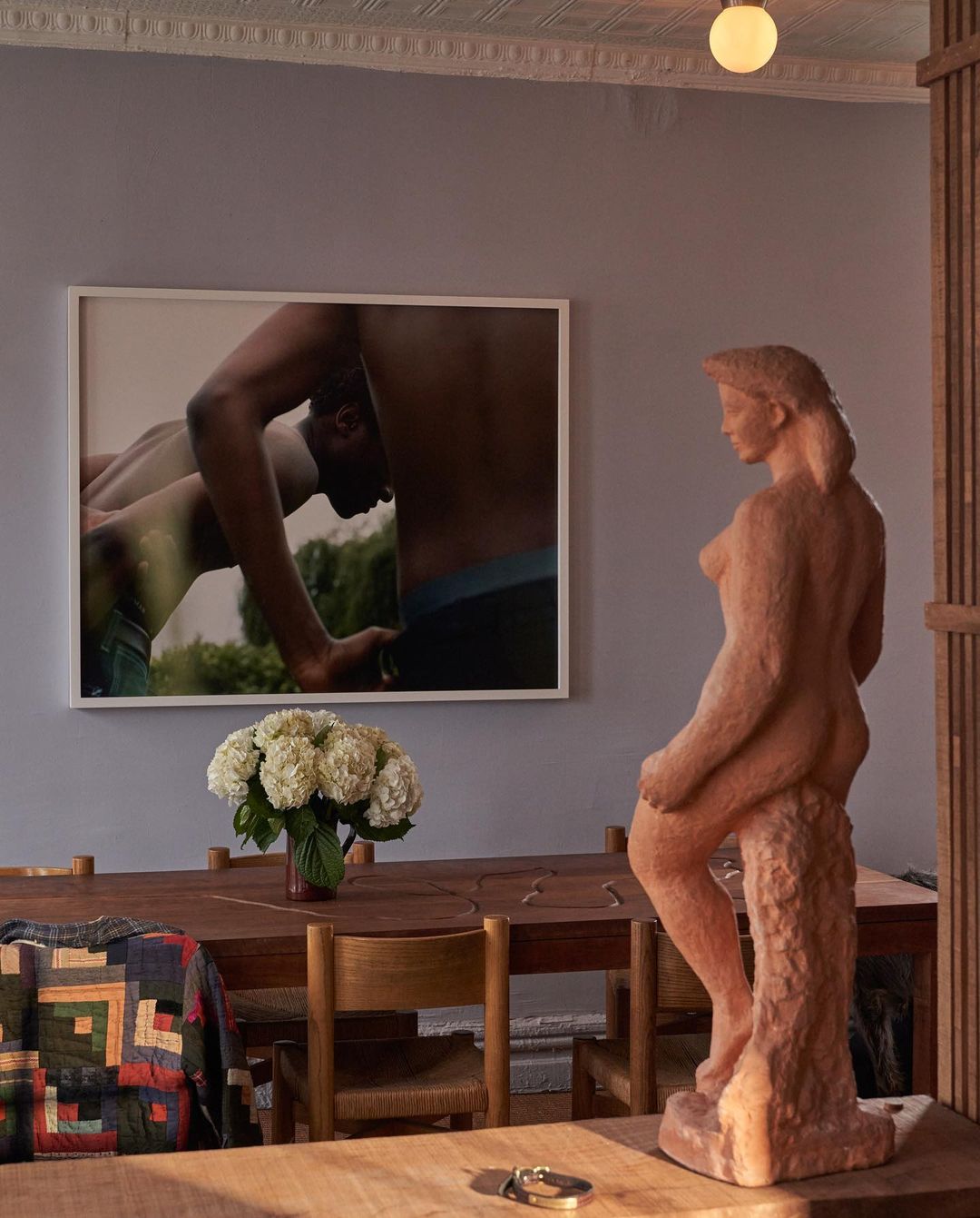 Interior shot with a nude sculpture on a table, floral arrangement, a framed art print of nude men, and natural light.