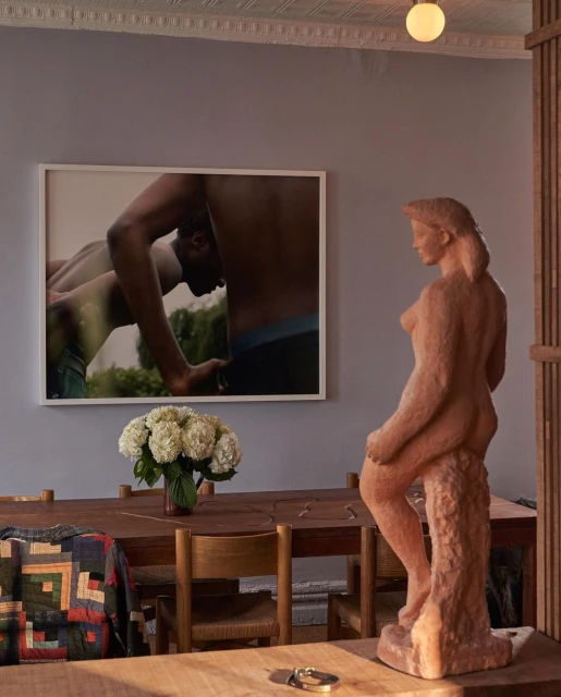Interior shot with a nude sculpture on a table, floral arrangement, a framed art print of nude men, and natural light.