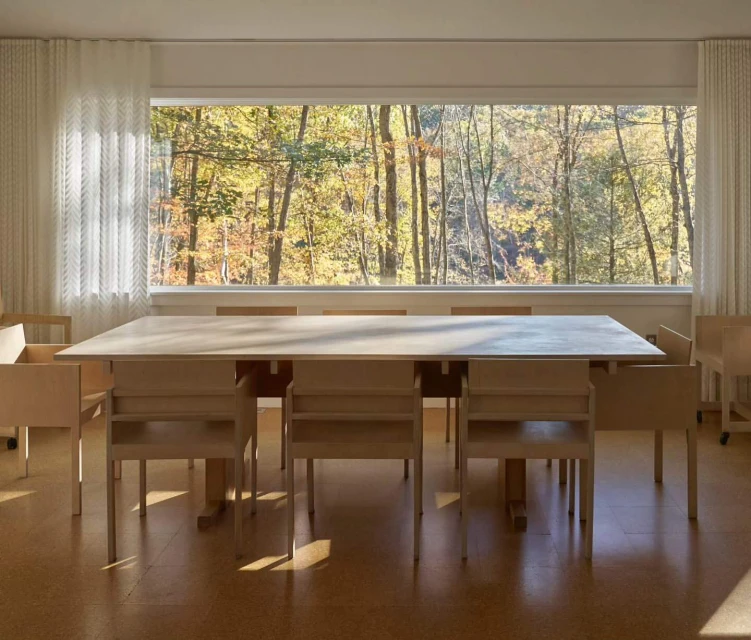 Interior view of a bright room with a light wood table and chairs. A large window overlooks a forest with colorful autumn foliage.
