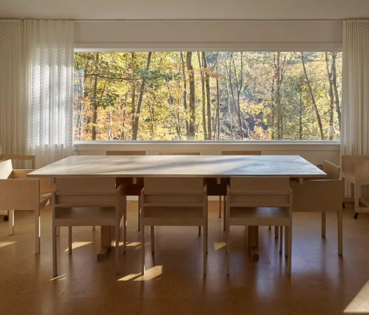Interior view of a bright room with a light wood table and chairs. A large window overlooks a forest with colorful autumn foliage.