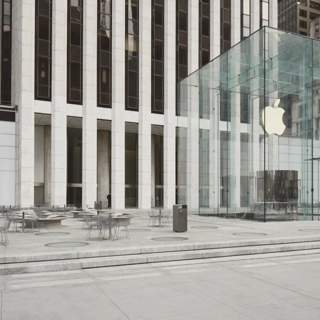 Exterior of Apple Store featuring the glass cube entrance, outdoor tables and chairs, and surrounding office buildings in an urban setting.