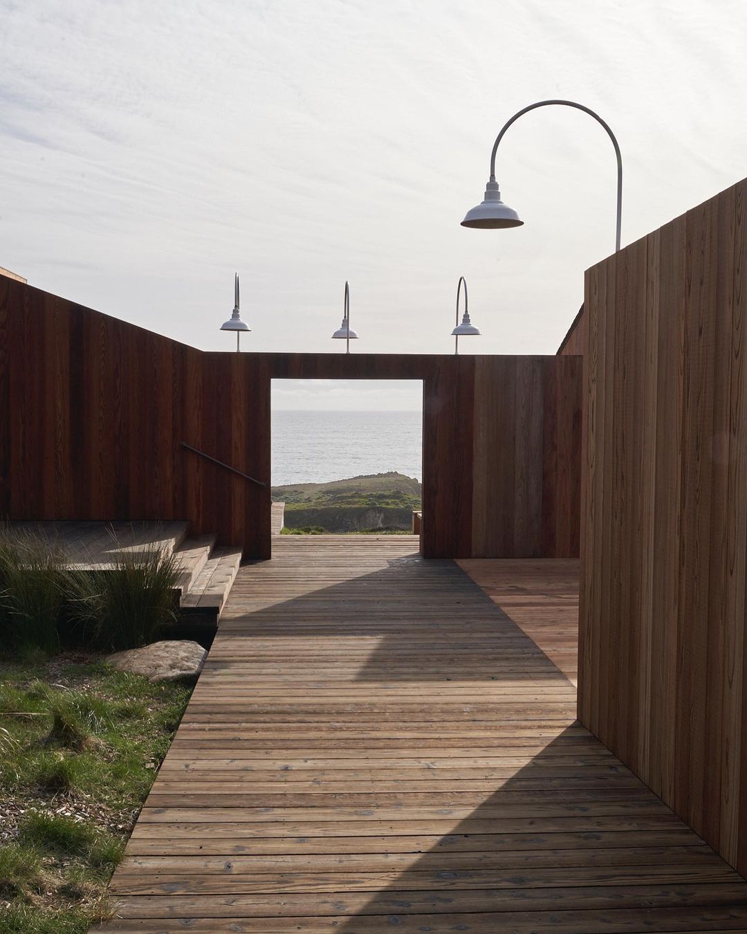 Wooden boardwalk leads to the ocean through an architectural structure with sconce lighting, creating a tranquil pathway.