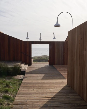 Wooden boardwalk leads to the ocean through an architectural structure with sconce lighting, creating a tranquil pathway.