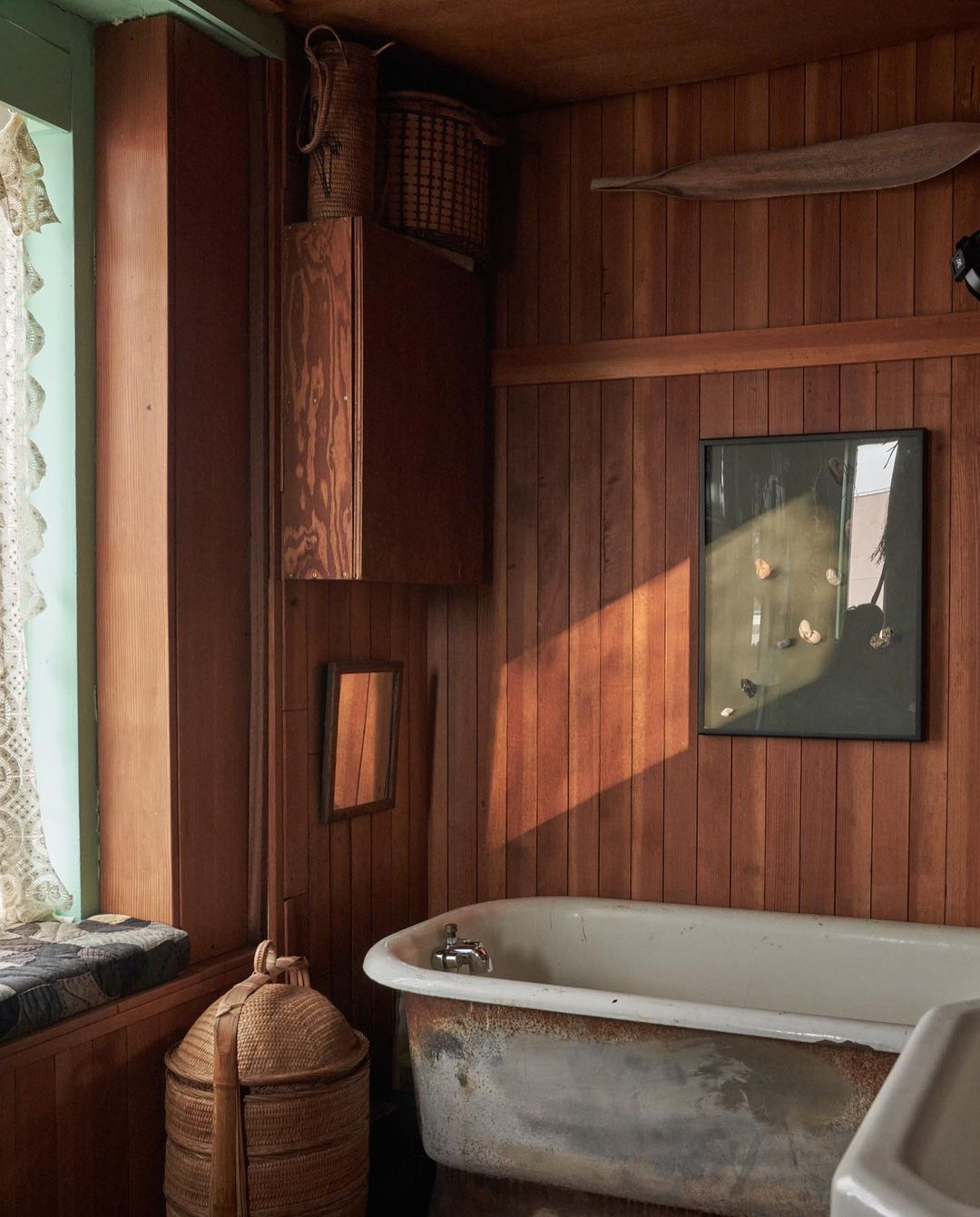 Rustic bathroom interior featuring a weathered bathtub, wood paneling, window seat, and woven baskets for a vintage aesthetic.