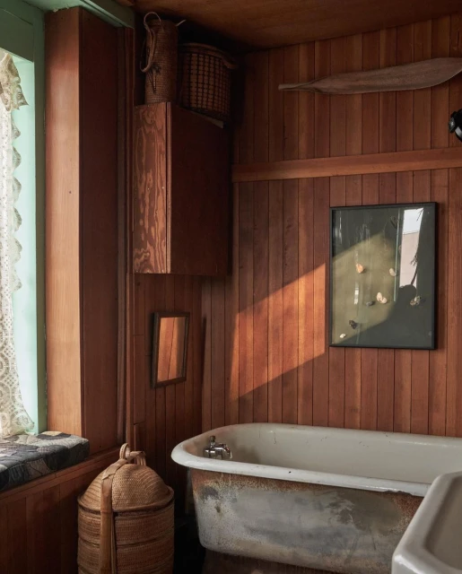 Rustic bathroom interior featuring a weathered bathtub, wood paneling, window seat, and woven baskets for a vintage aesthetic.