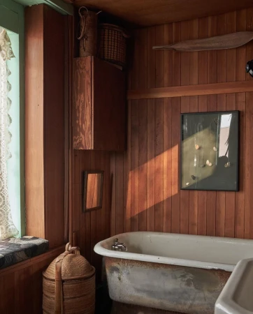 Rustic bathroom interior featuring a weathered bathtub, wood paneling, window seat, and woven baskets for a vintage aesthetic.