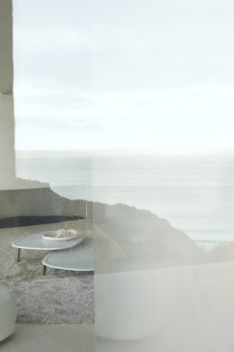 Minimalist home interior view of a hillside with ocean view, light grey color palette, two triangle-shaped coffee tables, and soft natural light.
