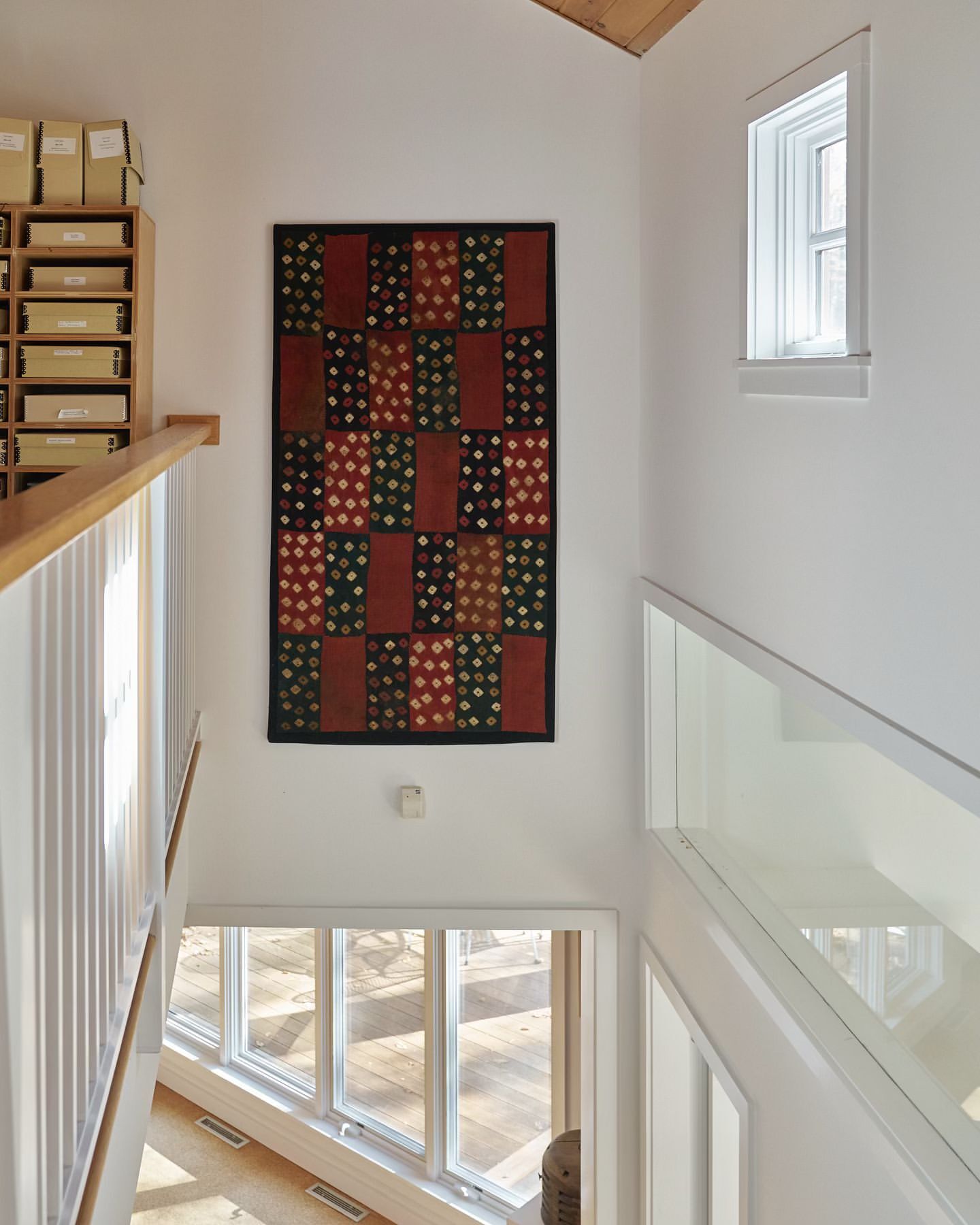 Interior view of a bright, modern home with a multi-paned window, wooden floors, white walls and staircase, and a colorful patterned wall hanging.