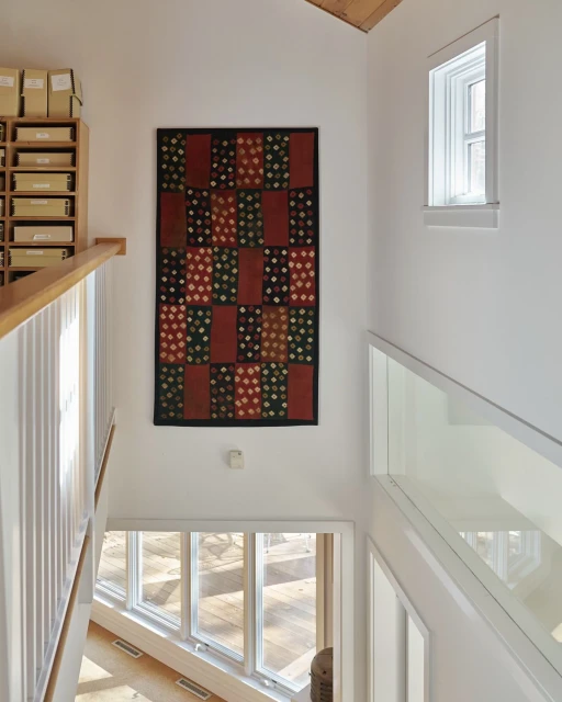 Interior view of a bright, modern home with a multi-paned window, wooden floors, white walls and staircase, and a colorful patterned wall hanging.