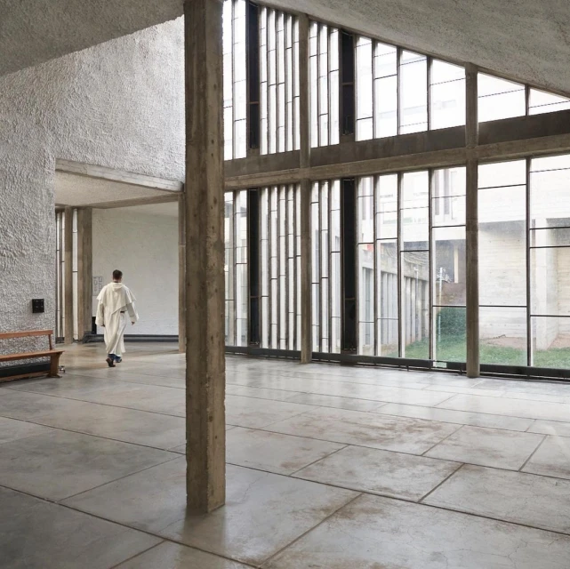 Interior view of a modern building with large windows and a person in a robe walking in the background. Concrete floor and column visible.