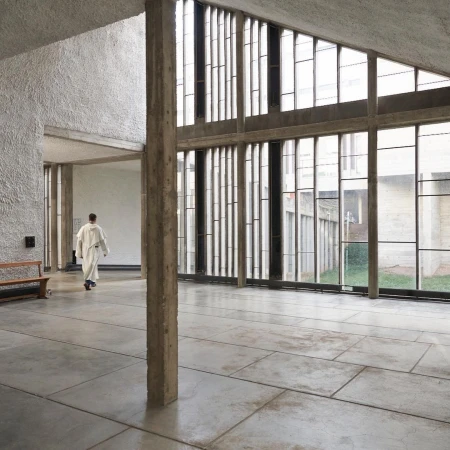 Interior view of a modern building with large windows and a person in a robe walking in the background. Concrete floor and column visible.
