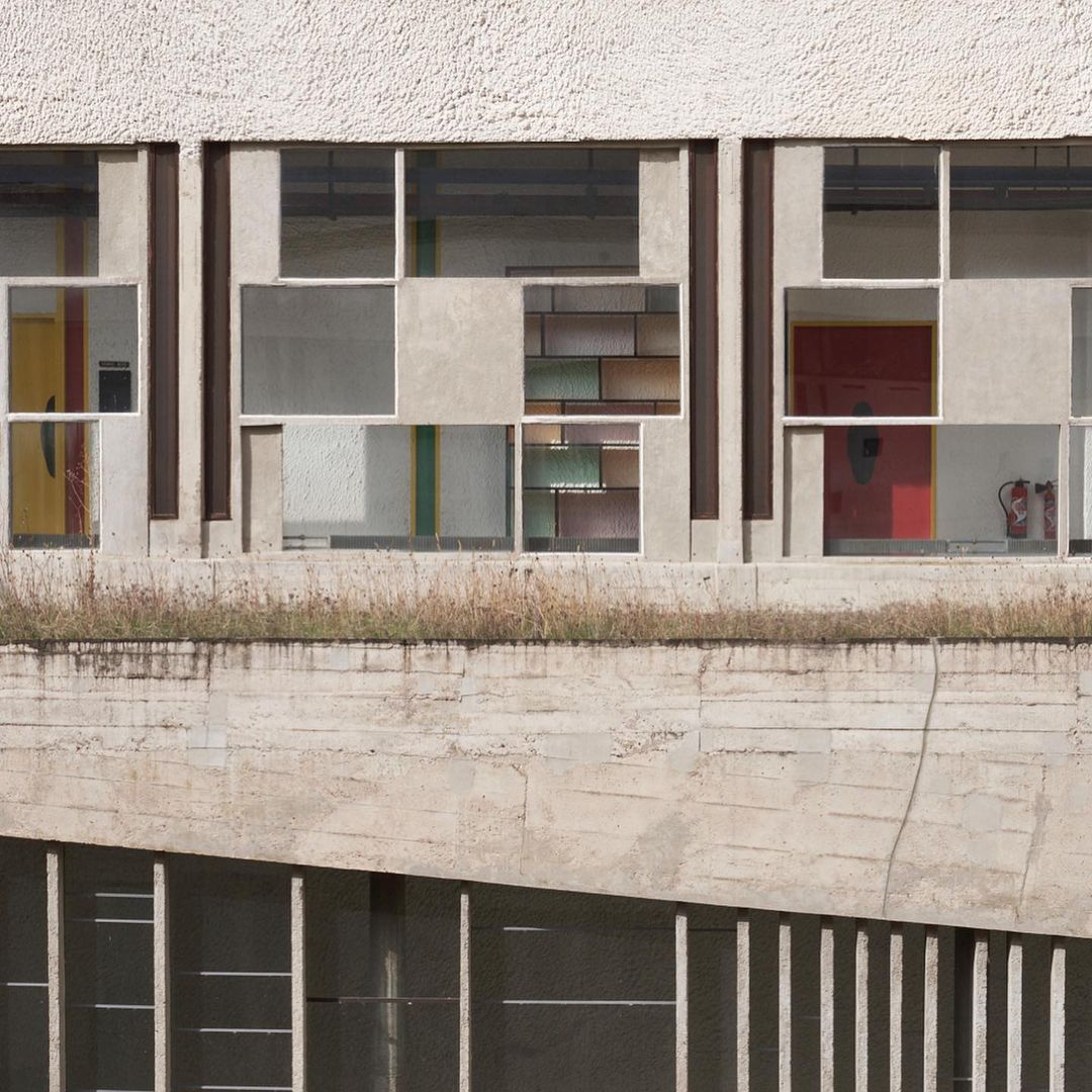 Exterior of modern architectural building featuring textured concrete, multi-paneled windows, and a flat roof with overgrown grass.