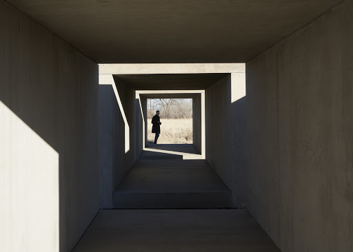 A person standing inside a rectangular concrete tunnel framing a view of a grassy field, captured by Martien Mulder.