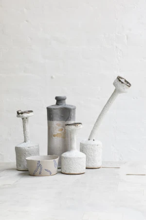 Still life featuring a collection of ceramic vases and a bowl with a leaf design, set against a textured white background.
