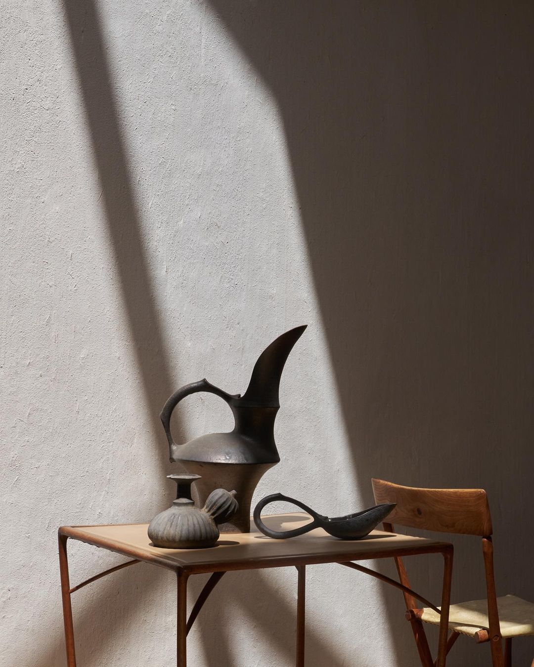 Still life of antique metal pitcher, vase, and spoon on a table next to a wood chair, against a textured wall with shadow.