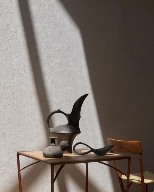 Still life of antique metal pitcher, vase, and spoon on a table next to a wood chair, against a textured wall with shadow.