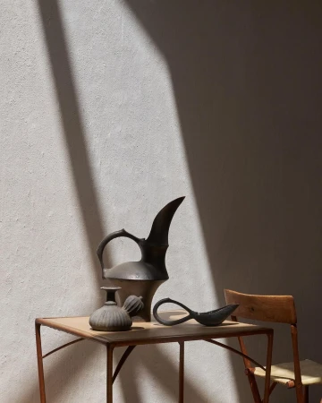 Still life of antique metal pitcher, vase, and spoon on a table next to a wood chair, against a textured wall with shadow.
