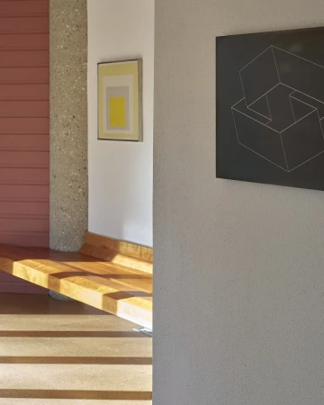 Interior architectural view showing wooden bench, concrete pillar, and framed abstract art on white and pink walls