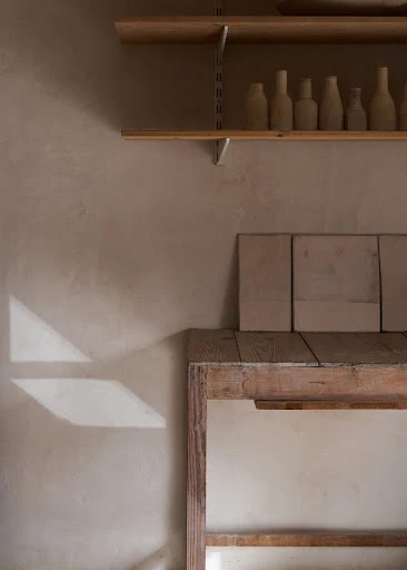 Minimalist photo of pottery display in neutral tones featuring shelves with clay bottles, square tiles, and geometric light patterns on wall.