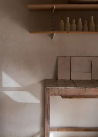 Minimalist photo of pottery display in neutral tones featuring shelves with clay bottles, square tiles, and geometric light patterns on wall.