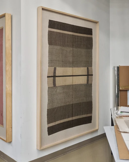 Framed textile art hangs on a white wall. The textile features horizontal bands in shades of brown and beige, with a linear pattern.