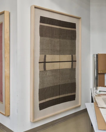 Framed textile art hangs on a white wall. The textile features horizontal bands in shades of brown and beige, with a linear pattern.