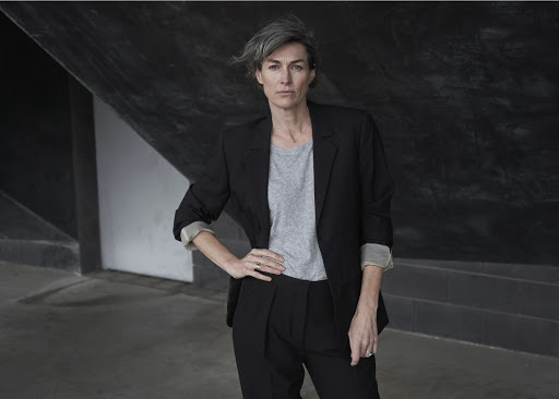 Professional portrait of a woman with short grey hair wearing a black suit and grey shirt, posing in an urban outdoor environment.