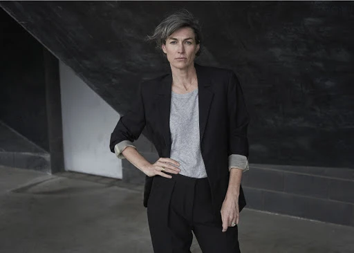 Professional portrait of a woman with short grey hair wearing a black suit and grey shirt, posing in an urban outdoor environment.