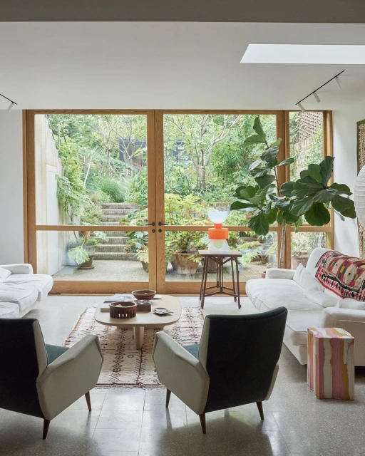 A bright living room with two white couches, two chairs, a coffee table, and a view of a lush green garden with stone stairs outside.