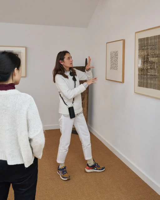 Two women at an art gallery look at framed artwork on a white wall, with a wood ladder visible in background, in natural lighting.
