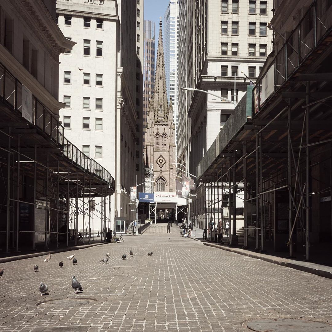 Urban shot of Trinity Church framed by skyscrapers in New York City, viewed down a street with brick pavement and scaffolding.