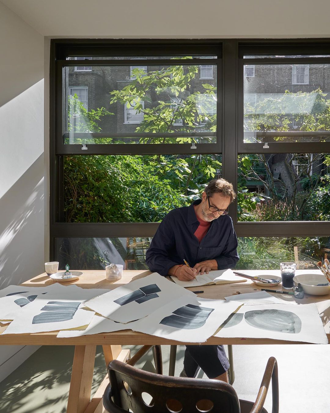 Adult male artist at table in studio by window, painting on paper, wearing glasses and a navy shirt, with green foliage outside.