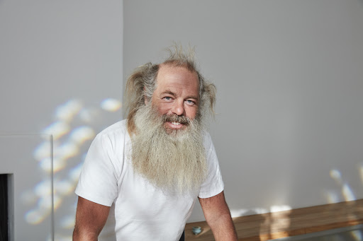Close-up portrait of a smiling adult male with a long white beard and messy hair, wearing a white t-shirt against a neutral background.