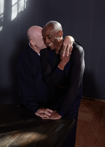 Two men embrace warmly indoors, one kissing the other's cheek; the foreground features a dark table, with natural light illuminating the scene.