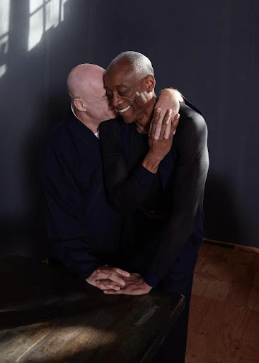 Two men embrace warmly indoors, one kissing the other's cheek; the foreground features a dark table, with natural light illuminating the scene.
