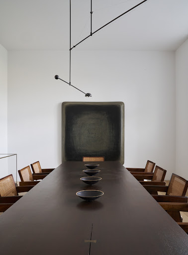Long brown conference table surrounded by wooden chairs in a bright, minimalist interior. A black painting hangs on a white wall.