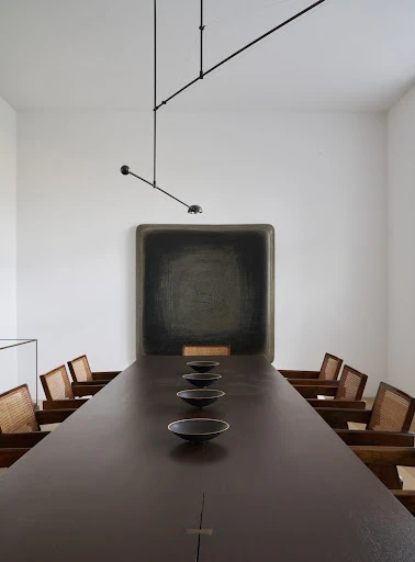 Long brown conference table surrounded by wooden chairs in a bright, minimalist interior. A black painting hangs on a white wall.