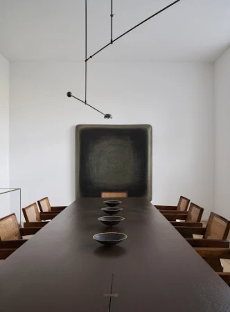 Long brown conference table surrounded by wooden chairs in a bright, minimalist interior. A black painting hangs on a white wall.