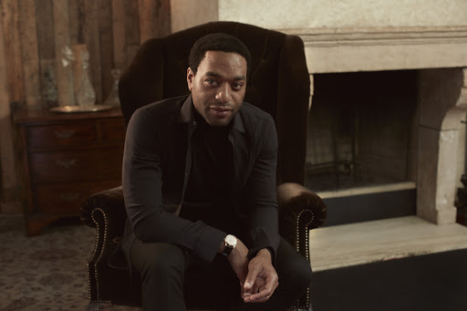 Elegant portrait of a seated adult male indoors. He wears a dark turtleneck and slacks, complemented by a wristwatch.