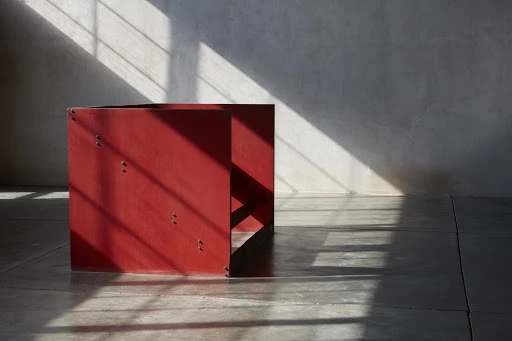 Abstract shot of geometric red box casting shadows on a concrete floor in front of a concrete wall, illuminated by sunlight.