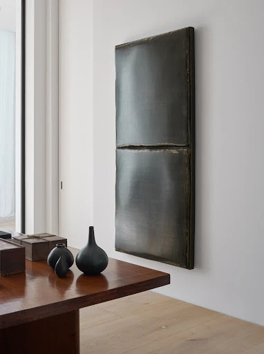 Abstract rectangular wall art hangs above a dark wood table with vases in a naturally lit interior space, showing home decor.