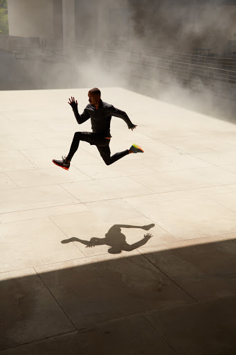 Athletic man mid-air, running in sportswear on urban plaza with stairs and diffused sunlight, creating a dynamic shadow silhouette.