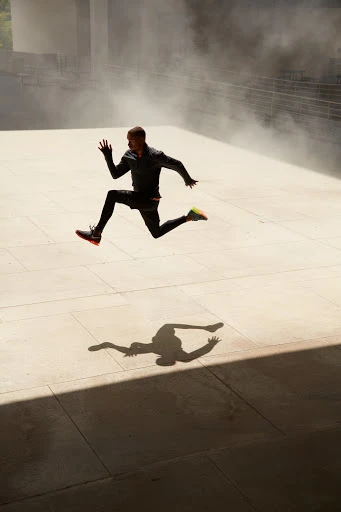 Athletic man mid-air, running in sportswear on urban plaza with stairs and diffused sunlight, creating a dynamic shadow silhouette.
