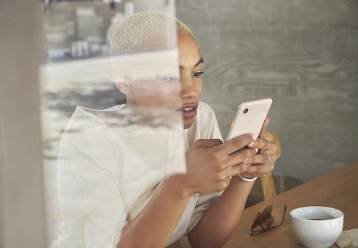 Woman with short blonde hair using a cell phone indoors at a table with a cup of coffee and glasses nearby, medium shot.
