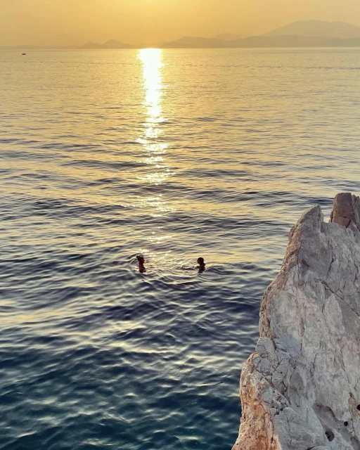 Two people swim in the ocean at sunset, viewed from a rocky cliff, with golden light reflecting on the water and distant land visible.