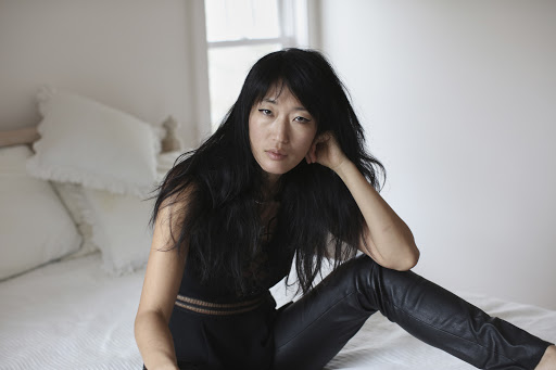 Adult woman with long black hair poses in dark top and leather pants, sitting on a bed in a softly lit indoor setting.