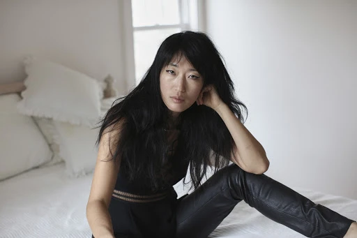 Adult woman with long black hair poses in dark top and leather pants, sitting on a bed in a softly lit indoor setting.
